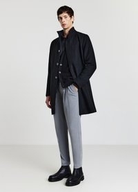 Schwarzer Wollmantel mit hohem Kragen und Knopfverschluss, getragen über einem schwarzen Pullover, kombiniert mit grauen maßgeschneiderten Hosen und schwarzen Stiefeletten.