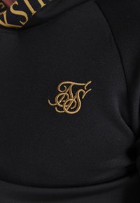 Camisa deportiva negra con una textura suave y tela perforada. Presenta un logo bordado en oro en el pecho y un diseño de cuello en contraste.