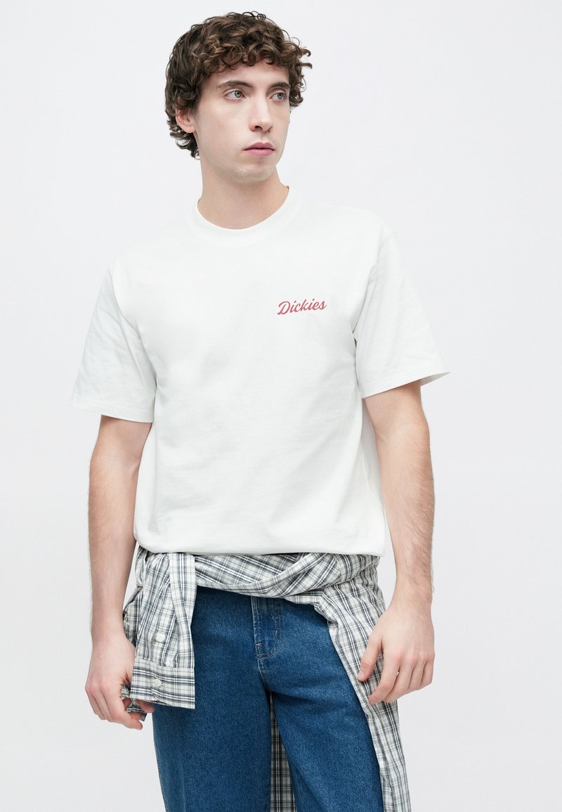 White cotton t-shirt featuring a red “Dickies” logo on the chest, paired with blue denim jeans and a plaid shirt tied around the waist.