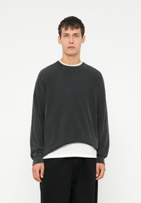 Dunkelgrauer, übergroßer Pullover mit Rundhalsausschnitt, aus weichem Stoff. Lange Ärmel und ein subtiler abgerundeter Saum. Über einem weißen T-Shirt getragen.