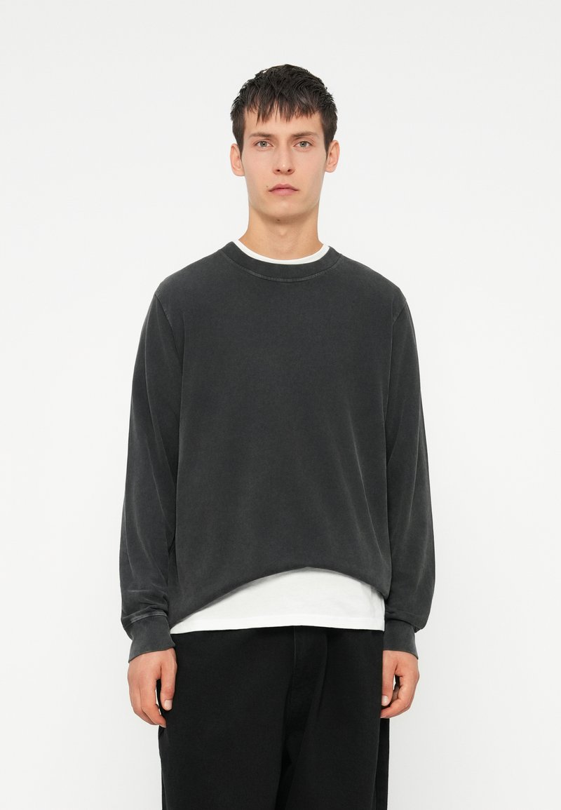 Dunkelgrauer, übergroßer Pullover mit Rundhalsausschnitt, aus weichem Stoff. Lange Ärmel und ein subtiler abgerundeter Saum. Über einem weißen T-Shirt getragen.