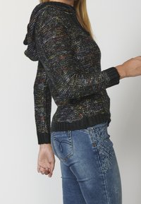 Černý pletený svetr s kapucí, zdobený barevnými skvrnami, s žebrovanými manžetami a lemem. Kombinován s modrými džínami s květinovou výšivkou po straně.