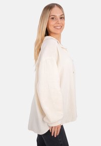 Helles beige Kapuzenshirt aus strukturiertem Stoff mit lockerer Passform. Hat eine Knopfleiste und breite Ärmel. Kombiniert mit dunklen Jeans.