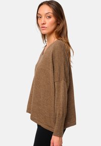 Femme aux cheveux châtain clair portant un pull brun à col en V ample et un pantalon noir, se tenant de profil contre un fond blanc.