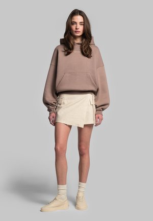 Felpa capuccio marrone con tasca frontale, abbinata a una gonna avvolgente beige con tasche laterali e cordini, e sneakers beige con calze di maglia.