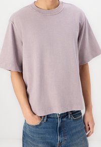 Camiseta de manga corta color lavanda, hecha de tela suave, con cuello redondo, corte holgado y detalles de costura visibles. Combinada con denim.