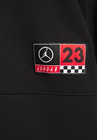 Tela negra con un parche rectangular cosido que presenta un logo Jumpman blanco, el número rojo "23", un patrón de tablero de ajedrez y el texto "JORDAN" en blanco.