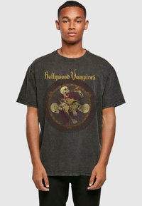 Juodas didelis t-shirt iš medvilnės, kuriame yra grafikų spausdinimas su kaukolėmis ir užrašas "Hollywood Vampires" auksine spalva priekyje.