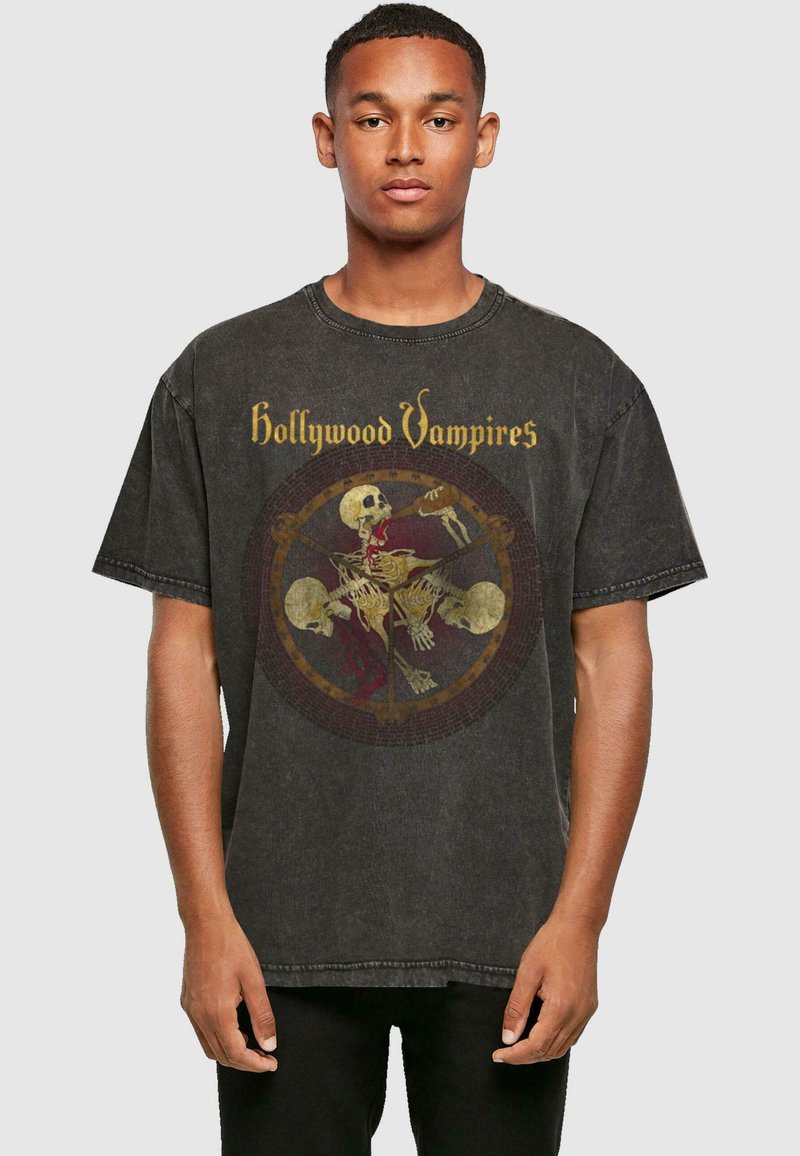 Juodas didelis t-shirt iš medvilnės, kuriame yra grafikų spausdinimas su kaukolėmis ir užrašas "Hollywood Vampires" auksine spalva priekyje.