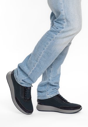 Person trägt hellblaue Jeans und schwarze Sneakers mit grauen Sohlen, steht mit einem leicht angehobenen Fuß vor weißem Hintergrund.