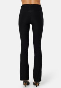 Zwarte flared broek van gladde stof, met een middelhoge taille, achterzakken en gestikte details. Gdragen met hoge hakken.