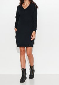 Schwarzes Sweatshirtkleid mit V-Ausschnitt, langen Ärmeln, Seitentaschen und einem goldenen Akzentknopf am Saum, kombiniert mit schwarzen Schnürstiefeln.