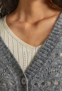 Grå stickad cardigan med ett texturerat mönster, knappstängning, lagd över en krämfärgad ribbad topp med V-hals och små ögla-detaljer.