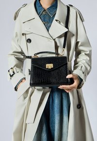 Borsa nera in finta pelle di coccodrillo con chiusura dorata, design strutturato, manico corto e tracolla removibile, appoggiata contro un trench coat chiaro.