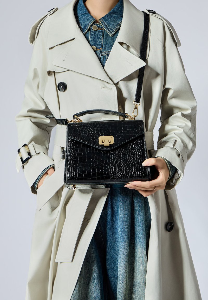 Borsa nera in finta pelle di coccodrillo con chiusura dorata, design strutturato, manico corto e tracolla removibile, appoggiata contro un trench coat chiaro.