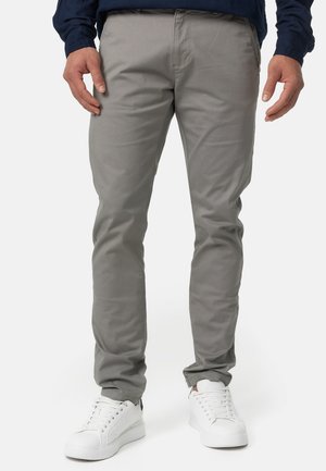 Homme portant un pantalon gris coupe slim et des baskets blanches, debout les mains détendues le long du corps devant un fond uni.