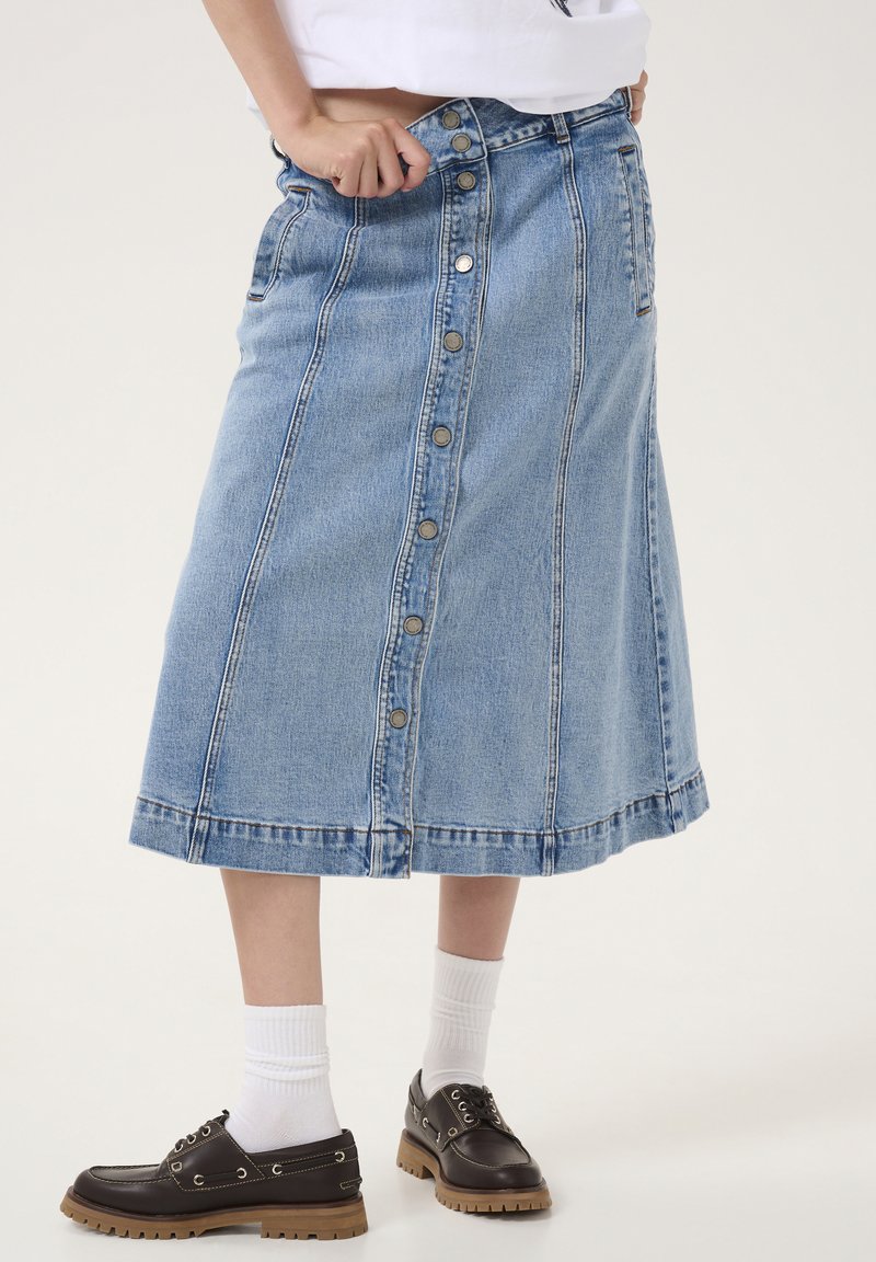 Denim midi krilo v svetlo modri barvi z zapiranjem na gumb spredaj, stranskimi žepi in razširjenim robom. Skombinirano s temno rjavimi čevlji in belimi nogavicami.