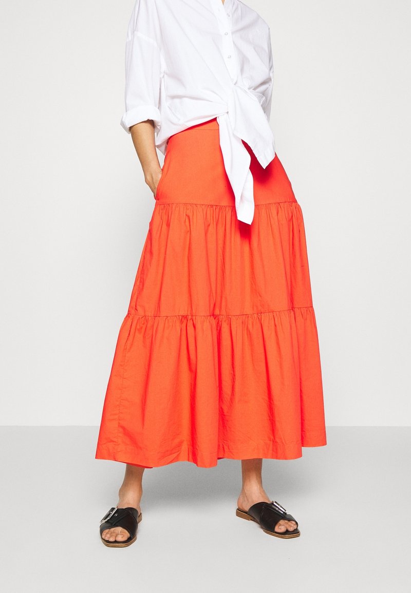 Nainen, jolla on päällään pitkä, kirkkaan oranssi kerroksellinen hame, valkoinen solmittu paita, jossa on käännetyt hihat, ja mustat avonaiset sandaalit, seisoo yksinkertaista taustaa vasten.