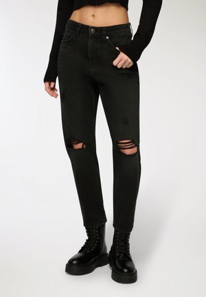 Jeans in denim nero con vita media, caratterizzati da tagli strappati sulle ginocchia e una vestibilità slim. Abbinati a stivali militari neri con lacci.