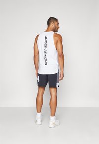 Bjela majica bez rukava s natpisom "UNDER ARMOUR" otisnutim okomito u crnoj boji na leđima. U kombinaciji s tamnim šortsevima i bijelim atletskim tenisicama.