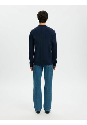 Mand står med ryggen til, iført en navyblå ribstrikket langærmet sweater og løstsiddende blå jeans med sorte sko på hvid baggrund.