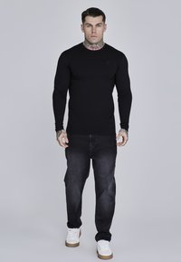 Crna majica dugih rukava s raglan rukavima, u kombinaciji s tamnim trapericama. Bijele tenisice i vidljive tetovaže na rukama i vratu.