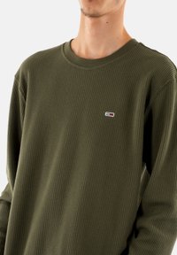 Camisa de manga larga verde oliva con un patrón de waffle texturizado. Presenta un pequeño logo multicolor en el área del pecho izquierdo.