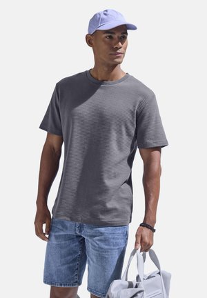 Mann mit lavendelfarbener Mütze, grauem strukturiertem T-Shirt, Jeans-Shorts und einer hellgrauen Tragetasche vor einfarbigem Hintergrund.