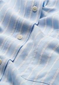 Chemise tissée bleu clair avec des rayures horizontales blanches et rouges, dotée de boutons blancs et de détails de couture visibles. Tissu lisse.