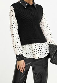 Top senza maniche a coste nere sovrapposto a una camicia bianca con motivi a cuore neri e finiture nere sui polsini, abbinato a pantaloni di pelle neri.