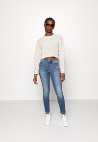 Maglione corto a maglia beige chiaro, jeans skinny blu con dettagli strappati e sneakers bianche. Modella con occhiali da sole scuri.