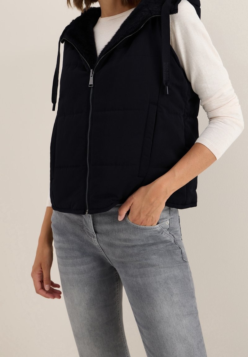 Femme portant un gilet sans manches noir matelassé avec fermeture éclair, chemise blanche à manches longues et jean gris, main dans la poche sur fond uni.