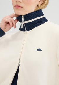 Chaqueta con cremallera de tejido crema con un alto cuello acanalado azul marino con rayas blancas. Pequeño logo en azul marino en el pecho. Textura suave.