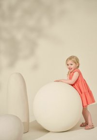 Weiße sphärische und elongated Objekte in einer hellbeigen Umgebung, mit einem Kind in einem korallenfarbenen Kleid, das sich an der Kugel lehnt.