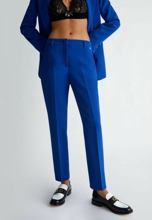 Pantalon bleu coupe slim associé à un soutien-gorge en dentelle noire, blazer bleu assorti et mocassins noirs et blancs sur un fond clair.