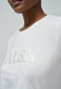 Camiseta de algodón blanca con escote redondo, que presenta un logo de lentejuelas plateadas texturizadas "SALSA JEANS" en la parte frontal.