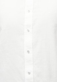 Μπροστινή όψη λευκού πουκάμισου με πέντε ορατά διαφανή κουμπιά και διακριτικό κεντητό λογότυπο "MK" στο επάνω δεξιό μέρος του στήθους.