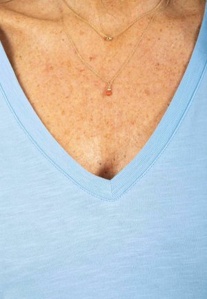 Jasnoniebieska bluzka z dekoltem w serek o miękkiej fakturze. Na szyi ułożone dwie złote naszyjniki, jeden z małym czerwonym wisiorkiem, leżące nad piegowatą obojczykiem.