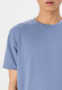 Τιραντέ μπλε μπλουζάκι με κοντά μανίκια που διαθέτει υφασμάτινο, γεωμετρικό μοτίβο. Στρογγυλή λαιμόκοψη και άνετη εφαρμογή. Μαλακό ύφασμα με διακριτές λεπτομέρειες.