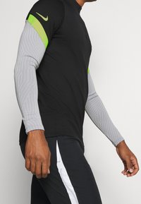 Schwarzes Sportshirt mit grauen langen Ärmeln, die ein strukturiertes Muster und leuchtend grüne Akzente auf den Schultern aufweisen. Nike-Logo ist vorhanden.