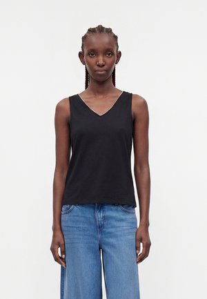 Femme aux cheveux tressés portant un haut noir sans manches à col en V et un jean bleu à jambes larges, debout devant un fond blanc uni.