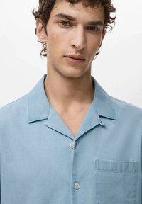 Camisa de manga corta de color azul claro con cuello de pico, cierre de botones en la parte frontal y bolsillo en el pecho izquierdo. Textura suave y corte casual.