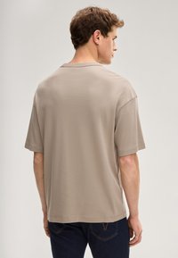 Beżowa bawełniana koszulka o luźnym kroju, z krótkimi rękawami i zaokrąglonym dekoltem. Widok z tyłu ukazuje minimalne oznakowanie marki i gładką fakturę.