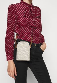 Bílá crossbody taška s monogramem, zlatými doplňky a řetízkem, kombinovaná s červenou vzorovanou blůzou a černými kalhotami.