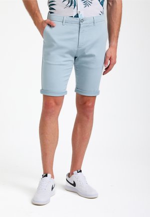 Pantalones cortos de algodón azul claro, con puños enrollados y bolsillos delanteros. Combinados con zapatillas blancas con detalles en negro. El fondo es blanco liso.