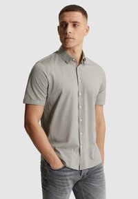 Homme aux cheveux courts portant une chemise gris clair à manches courtes boutonnée et un jean gris, mains dans les poches, regardant sur le côté.