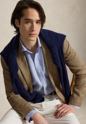 Jeune homme assis portant un blazer marron à carreaux, une chemise bleu clair, un pantalon blanc et un pull bleu marine posé sur les épaules.