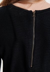 Pull noir avec une texture côtelée et un détail de fermeture éclair à l'arrière. La fermeture éclair est métallique et se prolonge verticalement le long de la couture arrière.
