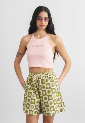 Γυναίκα με μακριά σκουρόχρωμα μαλλιά, που φοράει ροζ κοντό τοπ με τιράντες και πράσινα σορτς με σχέδια, με μωβ αποχρωματικά γυαλιά ηλίου και τατουάζ στο αριστερό μπράτσο.