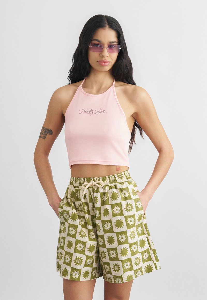 Γυναίκα με μακριά σκουρόχρωμα μαλλιά, που φοράει ροζ κοντό τοπ με τιράντες και πράσινα σορτς με σχέδια, με μωβ αποχρωματικά γυαλιά ηλίου και τατουάζ στο αριστερό μπράτσο.